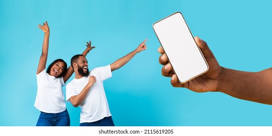Great Offer. Cheerful African American couple dancing and pointing at giant cell phone showing blank space for mock up, celebrating win, hand holding white empty screen, blue studio background wall