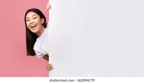 Great Offer. Cheerful Asian Girl Peeking Out Of Empty White Poster Advertising Something Over Pink Studio Background. Mockup, Panorama