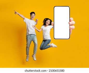 Great Offer. Excited Asian Couple Holding Big Smartphone With Empty White Cell Screen In Hand, Cheerful Guy And Lady Jumping Celebrating Win, Yellow Orange Studio Background, Mock Up Collage Banner