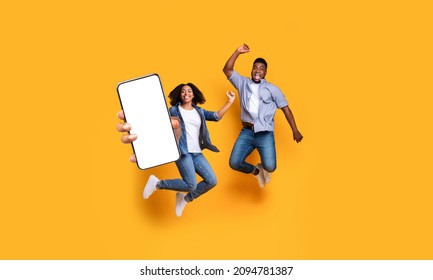 Great Offer. Excited Black Couple Holding Big Smartphone With Empty White Cell Screen In Hand, Cheerful Guy And Lady Jumping Celebrating Win, Yellow Orange Studio Background, Mock Up Collage Banner