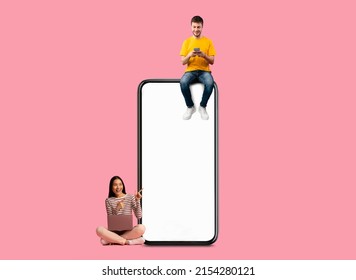 Great Offer. Excited Couple Sitting On Big Giant Smartphone Using Gadgets, Cheerful People Chatting On Social Media, Asian Female With Pc Pointing At Blank White Screen, Pink Wall, Mock Up Banner