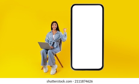 Great Offer. Excited lady sitting on chair using laptop pc pointing finger at big cellphone with empty white screen, presenting new cool app, free copy space mock up, website design banner - Shutterstock ID 2111874626