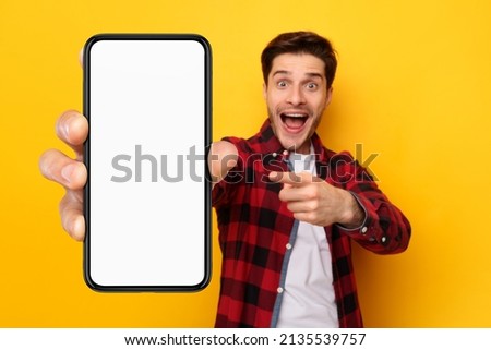 Great Offer. Excited surprised guy holding and pointing finger at big giant cell phone with white screen in hand, promoting application or website, advertising product or service, yellow studio wall