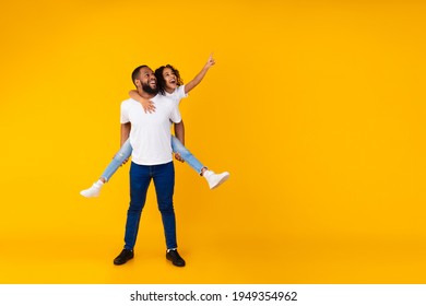 Great Offer. Full Body Length Of Cheerful African American Man Giving Piggy Back Ride For His Daughter, Little Girl Looking Aside And Pointing Fingers Up At Free Copy Space On Yellow Background