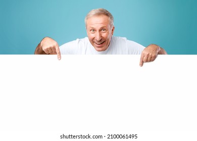 Great Offer. Happy Excited Mature Man Peeping Out Of White Advertisement Board, Pointing Fingers Down At Free Space For Your Text Or Design. Smiling Cheerful Guy Showing Mockup, Standing On Blue Wall