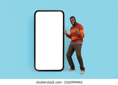 Great Offer. Overjoyed Black Man Celebrating Success Near Big Blank Smartphone With White Screen, Happy Young African American Male Clencing Fists And Exclaiming With Excitement, Mockup Image - Powered by Shutterstock