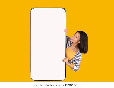 Great Offer. Portrait of excited asian woman peeking out big giant vertical cell phone with white blank screen and looking at device display. Gadget with empty free space mock up, yellow orange wall - Shutterstock ID 2119015955