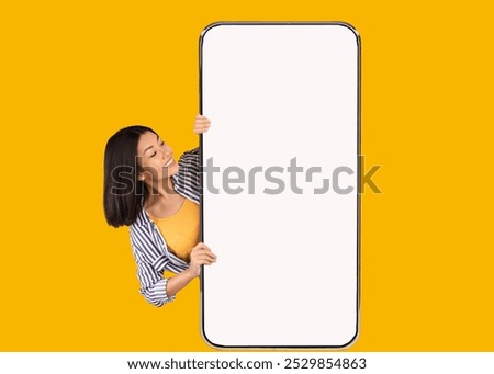 Great Offer. Portrait of excited asian woman peeking out big giant vertical cell phone with white blank screen and looking at device display. Gadget with empty free space mock up, yellow orange wall