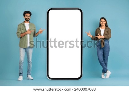Great offer for two. Cheerful young indian couple posing by big smartphone with white blank screen, showing newest mobile application, mockup, copy space, blue background
