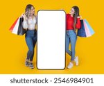 Great online shopping app, excited full body two young caucasian women standing near big mobile phone mock up with empty huge touch screen hold carry shopping bags isolated on yellow background.