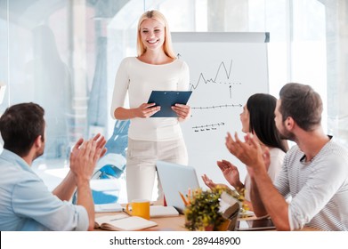 Great presentation! Cheerful young woman standing near whiteboard and smiling while her colleagues sitting at the desk and applauding