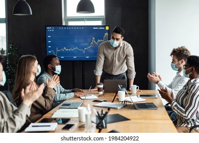 Great Presentation. Portrait Of Happy Arab Corporate Leader In Surgical Mask Giving Speech During Workshop In Modern Office, Standing In Boardroom, Diverse Teammates Sitting At Desk Clapping Hands