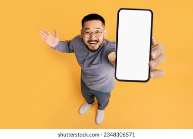 Great Promo. Excited Asian Man Showing Blank Smartphone With White Screen At Camera, Cheerful Guy Recommending New Mobile App Or Website, Standing Over Yellow Studio Background, Above Shot, Mockup