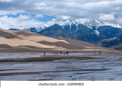 Great Sand Dunes National Park & Preserve, Colorado, USA - May 06, 2016: A Group Kids Are Playing In Medano Creek At The Base Of Sand Dunes And Snow-capped Peaks As Spring Clouds Passing Over.