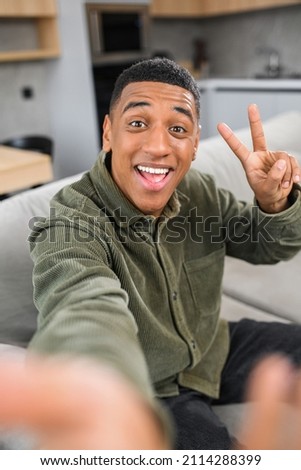 Great selfie. Multiracial young guy gesturing to the smartphone camera, while making funny photos at home at the sofa