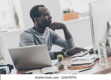 Great solutions every day. Handsome young African man keeping hand to his face while sitting at his working place in home office