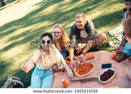 Great time with friends. Gheerful friends sitting on the grass taking selfie and eating pizza after doing their homework.
