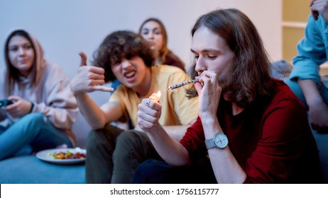 Great Time. Young Teenager, Man Lighting Cannabis Cigarette While Sitting With Excited Friends On The Couch At Home. Young People Smoking Marijuana Together, Having Fun. Marijuana, Weed Legalization