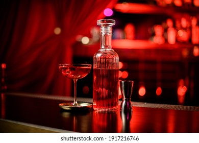 Great View Of Crystal Bottle With Liqueur And Glass Of Cocktail On Bar Counter. Blurred Background. Red Backlight