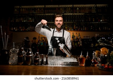 great view of smiling man bartender skillfully pouring ice cubes from metal scoop into shaker cup. Different bar equipment on the bar counter at foreground - Powered by Shutterstock