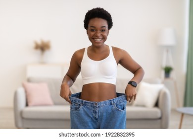 Great Weight Loss. Thin Black Lady Wearing Large Jeans Showing New Small Size And Result Of Successful Slimming Standing Posing At Home, Smiling To Camera. Weight-Loss Motivation Concept