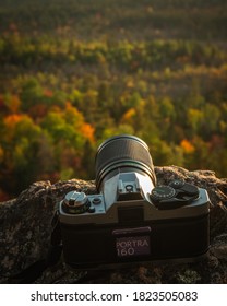 GREATER MADAWASKA, ONTARIO, CANADA - SEPTEMBER 27, 2020: A 35mm camera loaded with Kodak Portra 160 film sits on rock at the edge of a cliff, aimed at a forest of autumn-colored trees below.