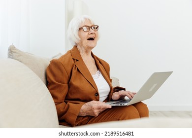 Greatly Surprised By The Shock, An Elderly Woman In A Brown Suit Is Sitting On A Beige Sofa With Her Mouth Wide Open And Holding A Laptop On Her Lap