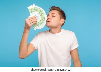 Greed and money. Young businessman in casual white t-shirt smelling euro bills with expression of pleasure, enjoying wealthy life, big money profit. indoor studio shot isolated on blue background