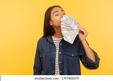 Greedy for money. Portrait of avaricious rich woman in denim shirt smelling earned dollar banknotes, enjoying success and big profit, wealthy life. indoor studio shot isolated on yellow background