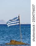 Greek flag waving on a small rock in the Aegean Sea, with blue waters and distant islands in the background, symbolizing Greek maritime heritage and natural beauty. High quality photo