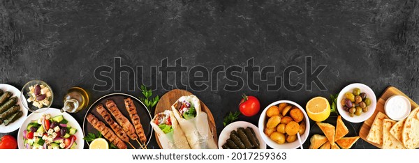 Greek food bottom border, top view on a dark\
banner background. Souvlaki, gyros wraps, salad, spanakopita,\
dolmades, pita and lemon potatoes. Copy\
space.