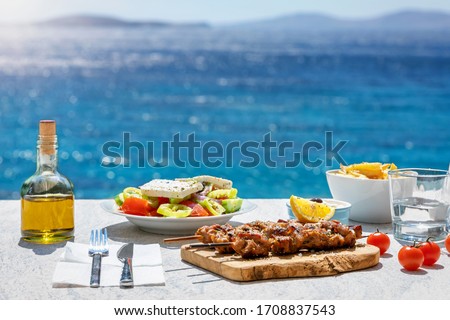 Greek food concept with farmers salad and souvlaki skewers in front of the sparkling, blue Aegean sea during summer time