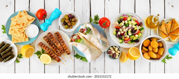 Greek food table scene, top view on a white wood banner background. Variety of items including gyros wraps, souvlaki, salad, spanakopita, dolmades, pita and lemon potatoes.