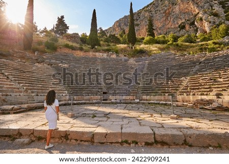 Greek history:  A long-haired woman from behind, wearing white dress  looking at Apollo Temple and Ancient Theater in sunset. Delphi, Greece.