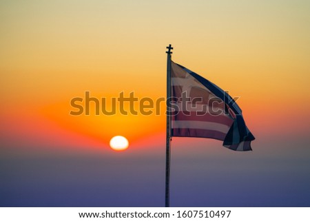 Greek national flag blowing in breeze back-lit by rising sun against colorful sky.