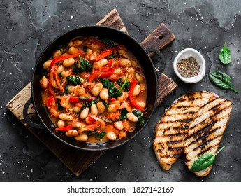Greek style tomato sauce, spinach, paprika, beans stew in a cast iron pan on a rustic board on a dark background, top view. Simple comfort food                    