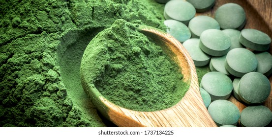 Green algae in powder and pills - chlorella, spirulina in wooden spoon on wooden background - closeup - banner design.