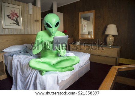 Green alien sitting using tablet computer on the bed in an old-fashioned room