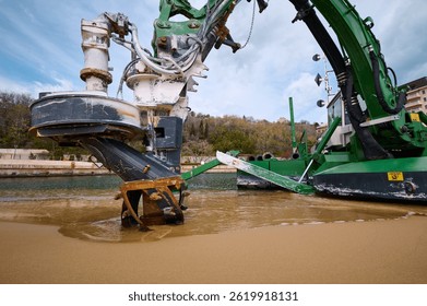 Green amphibious dredging machine working at the edge of a beach and water channel, used for underwater excavation and maintenance near a marina or coastal area