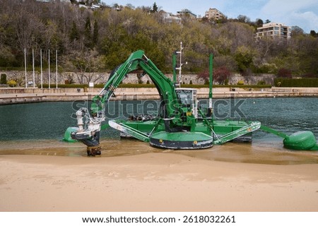 Green amphibious dredging machine working at the edge of a beach and water channel, used for underwater excavation and maintenance near a marina or coastal area