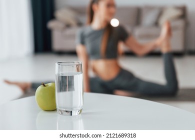 Green apple and glass of water is on the table. Young woman with slim body type and in yoga clothes is at home. - Shutterstock ID 2121689318