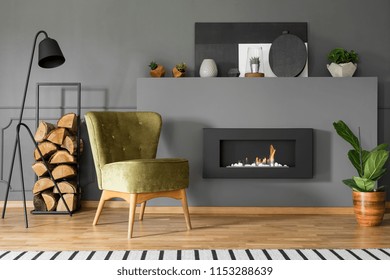 Green armchair and firewood next to fireplace in grey living room interior with lamp. Real photo