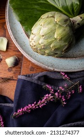 A Green Artichoke In Ceramics Plate Decorated With Beautiful Flower On Wooden Table And Cotton Fabric, Bright Lighting , Dramatic Food Photography