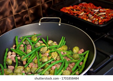 Green Beans And Brussel Sprouts With Bacon Bits In A Deep Pan On An Oven Top Together With A Roasting Dish With Red Peppers And Onions In The Background Against A Copper Tiled Backsplash.