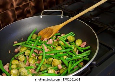 Green Beans And Brussel Sprouts With Bacon Bits In A Deep Pan On An Oven Top Against A Copper Tiled Backsplash.  A Wooden Spoon Rests In The Black Pan.