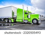 Green big rig classic American bonnet semi truck tractor with extended cab compartment for truck driver rest and dry van semi trailer standing on the industrial parking lot waiting for next freight