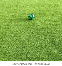 A green and black striped ball resembling a watermelon lies on a lush, well-maintained grassy field. The vibrant colors and simplicity make it visually appealing and suitable for various uses. - Powered by Shutterstock