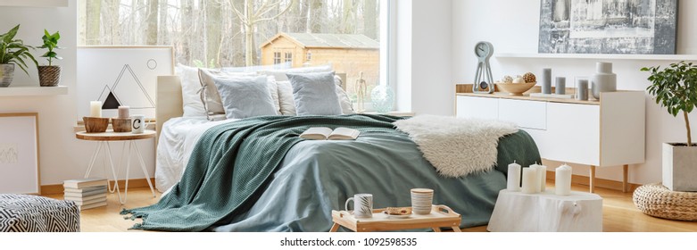 Green blanket on a cozy, double bed by a large window in a scandinavian style bedroom interior with a view at a forest - Shutterstock ID 1092598535