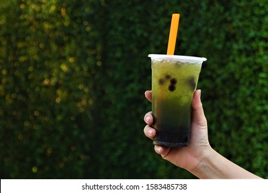 The Green Boba Tea In A Clear Plastic Cup With An Orange Straw Held In The Hand, With A Backdrop Of A Tree Wall.