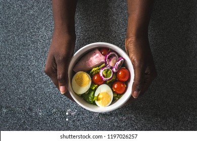 Green breakfast in bowl with spinach, cherry tomatoes, eggs, onions and red tomatoes. African Girl holding bowl with hands visible, top view. For Clean eating, dieting, weight loss food concept